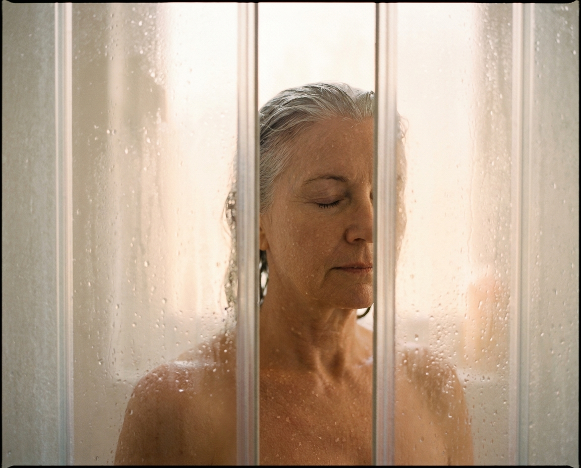 Silver-haired woman in her early 60s seen through frosted shower glass in warm steam, eyes closed, water droplets on fair skin