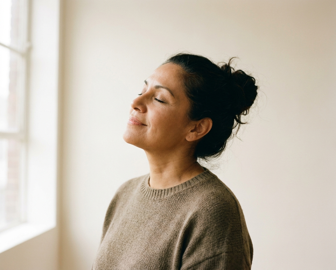 Latina woman in her late 40s with eyes closed and face tilted toward soft window light in a studio setting