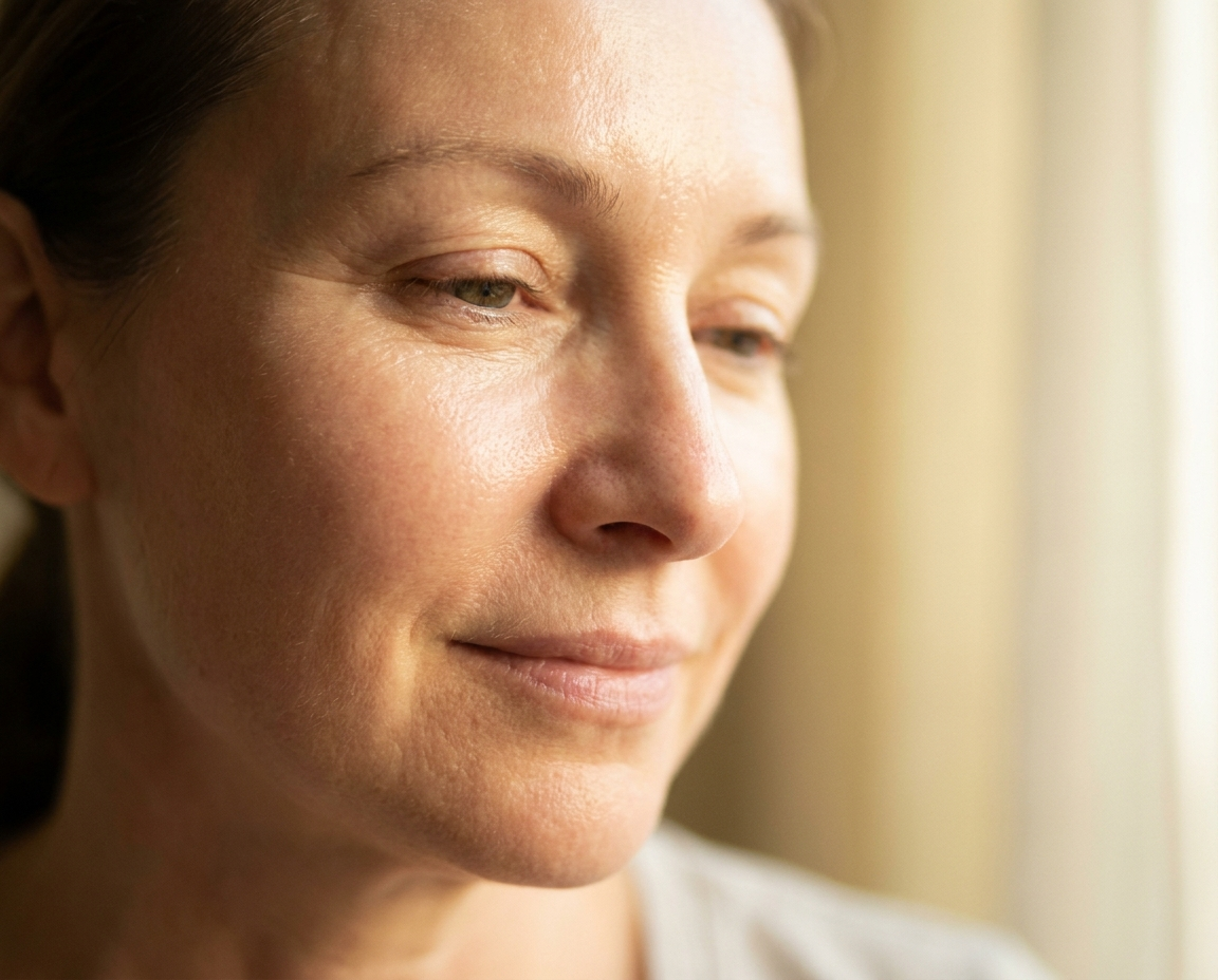 Close-up portrait of a woman in her early 50s with luminous post-facial skin, no makeup, soft warm light revealing incredible texture