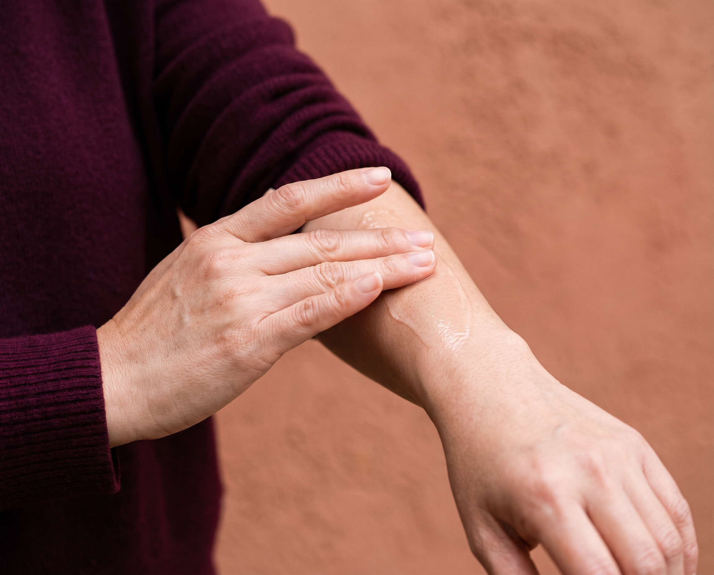 Asian woman's hands patting serum into forearm skin, wearing a deep burgundy cashmere sweater against a terracotta background