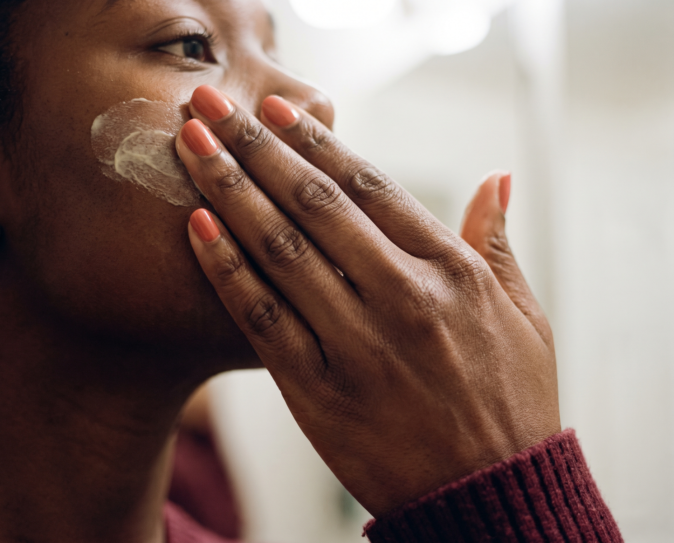 Close-up of a woman with deep brown skin smoothing cream cleanser across her cheek, coral nails and burgundy top