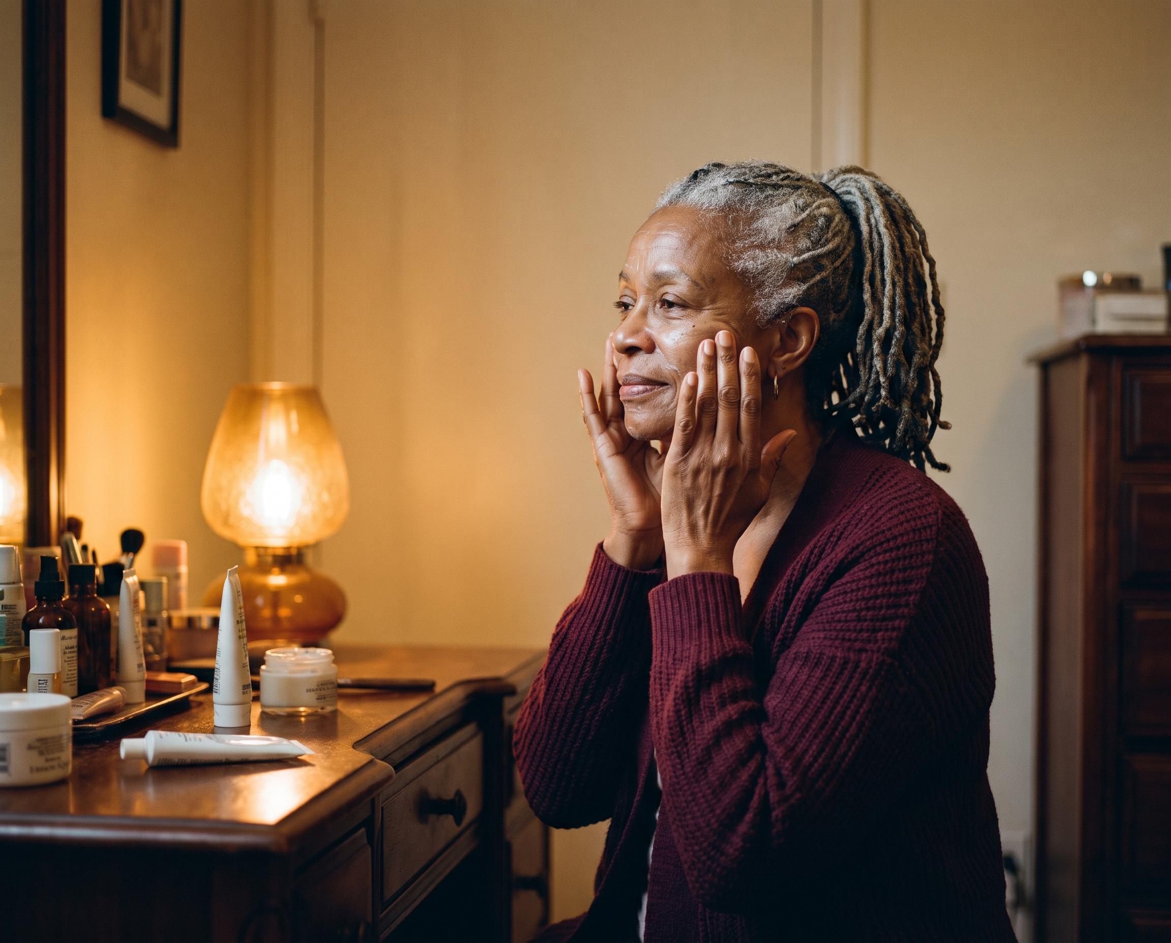 A Black woman in her 60s with silver locs applying moisturizer at a warm walnut vanity in amber lamp light