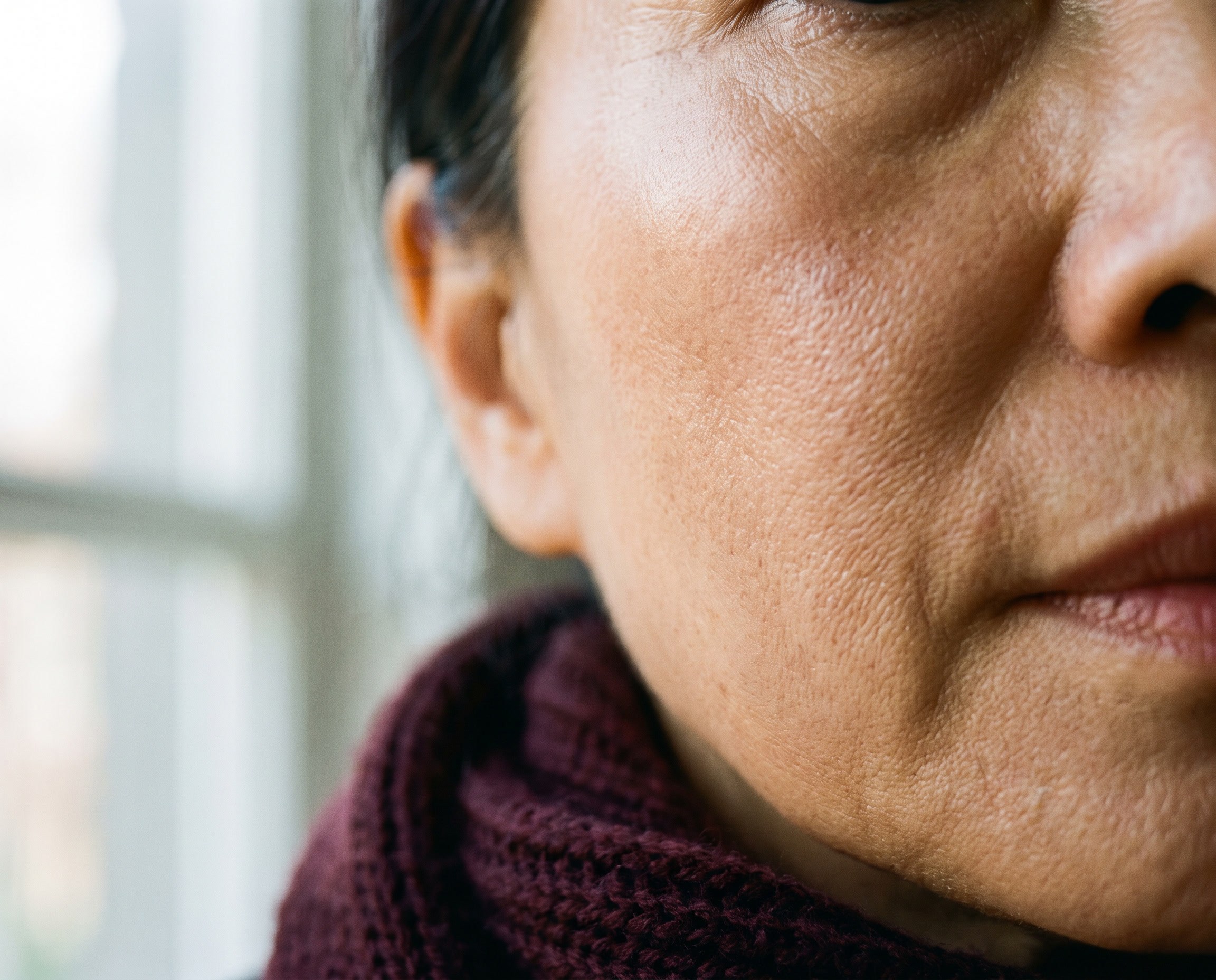 Close-up of glowing skin on the cheekbone of a woman in her early 60s of East Asian heritage, soft window light, burgundy scarf at frame edge