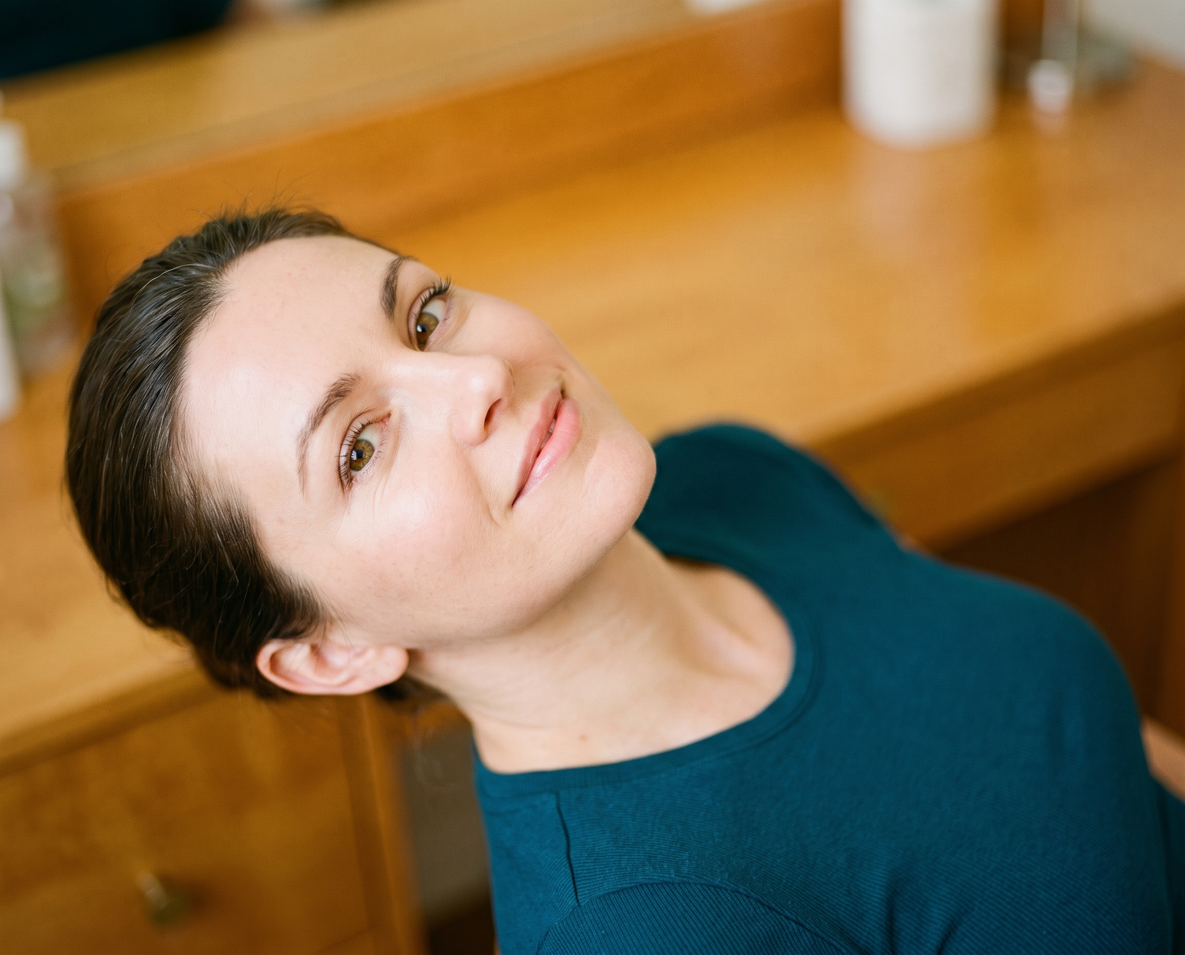 Close-up beauty shot of a fresh-faced woman in a deep teal top, head tilted back with bright eyes and smooth skin