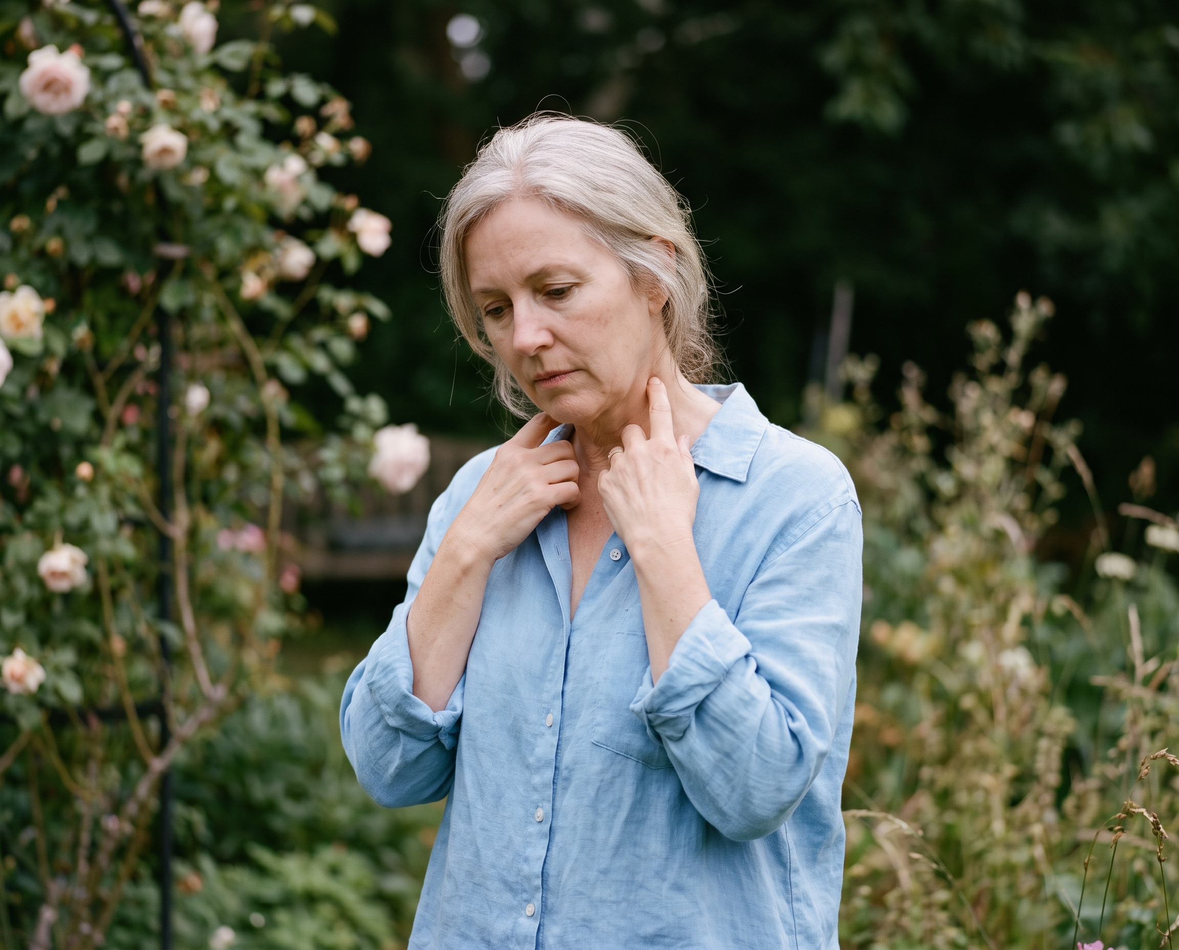 A woman in her late 50s outdoors in soft shade, gently touching her collarbone and neck, blurred garden in the background
