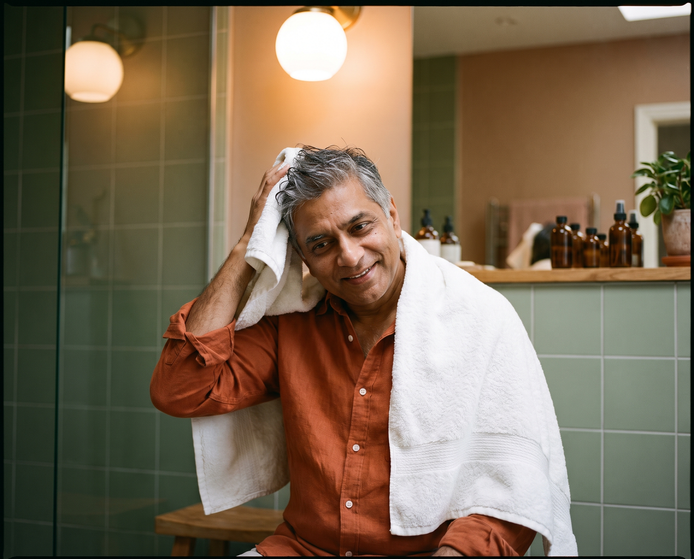 South Asian man in his late 50s in a rust-orange shirt towel-drying his hair in a sage-green bathroom