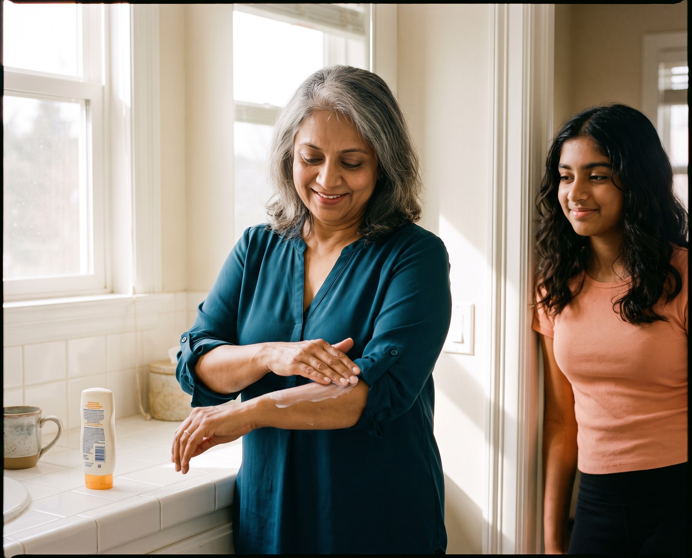 South Asian woman in her late 50s in a deep teal blouse applying sunscreen to her forearms at a bathroom sink while her teenage granddaughter watches