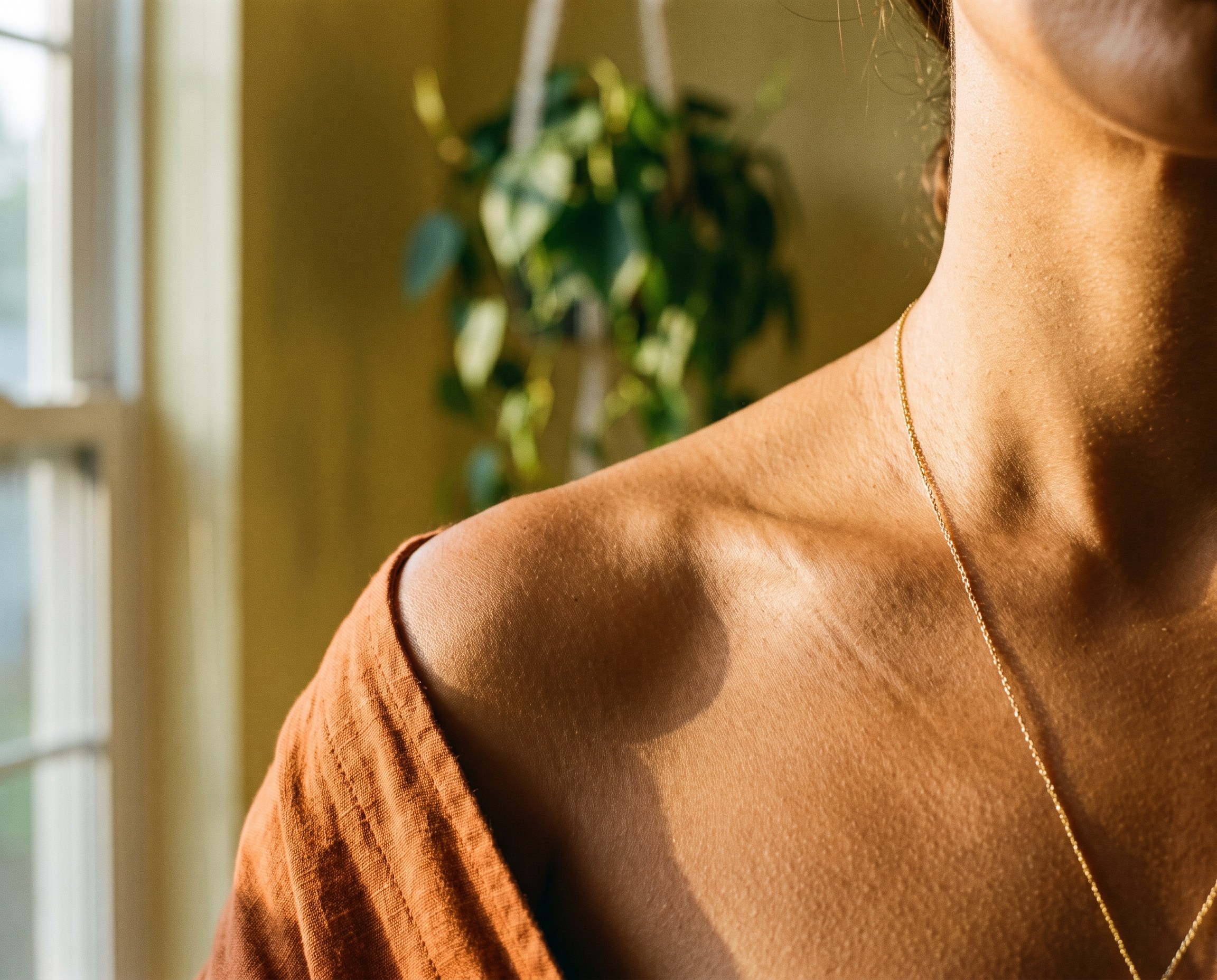 Close-up of warm honey-brown skin on a woman's collarbone with a gold chain, late-afternoon window light, rust-orange linen fabric at shoulder