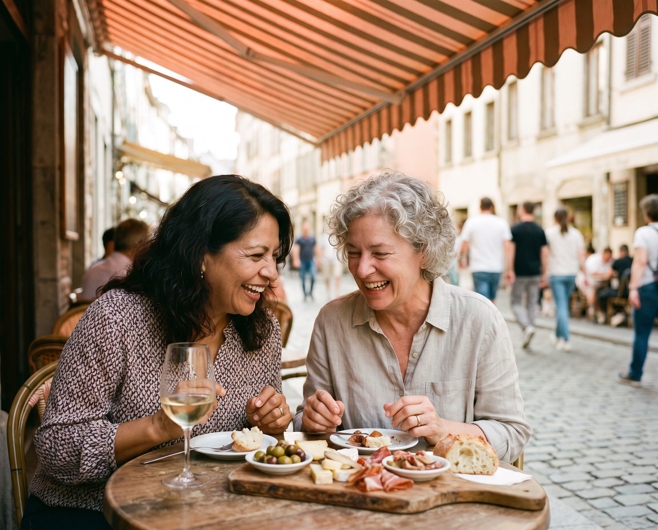 Two women in their late 50s and early 60s - one Latina, one white with curly gray hair - sharing tapas and laughing at an outdoor Mediterranean cafe