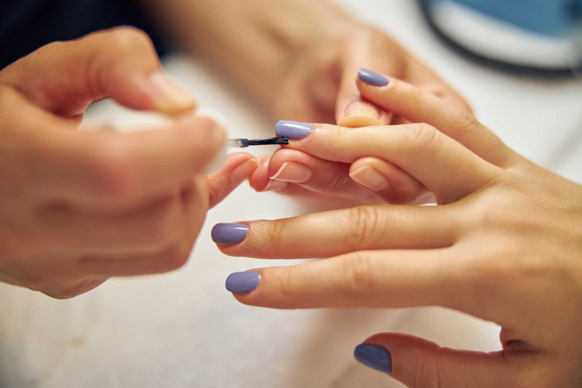 Manicurist applies lavender nail polish to a client’s fingernail.
