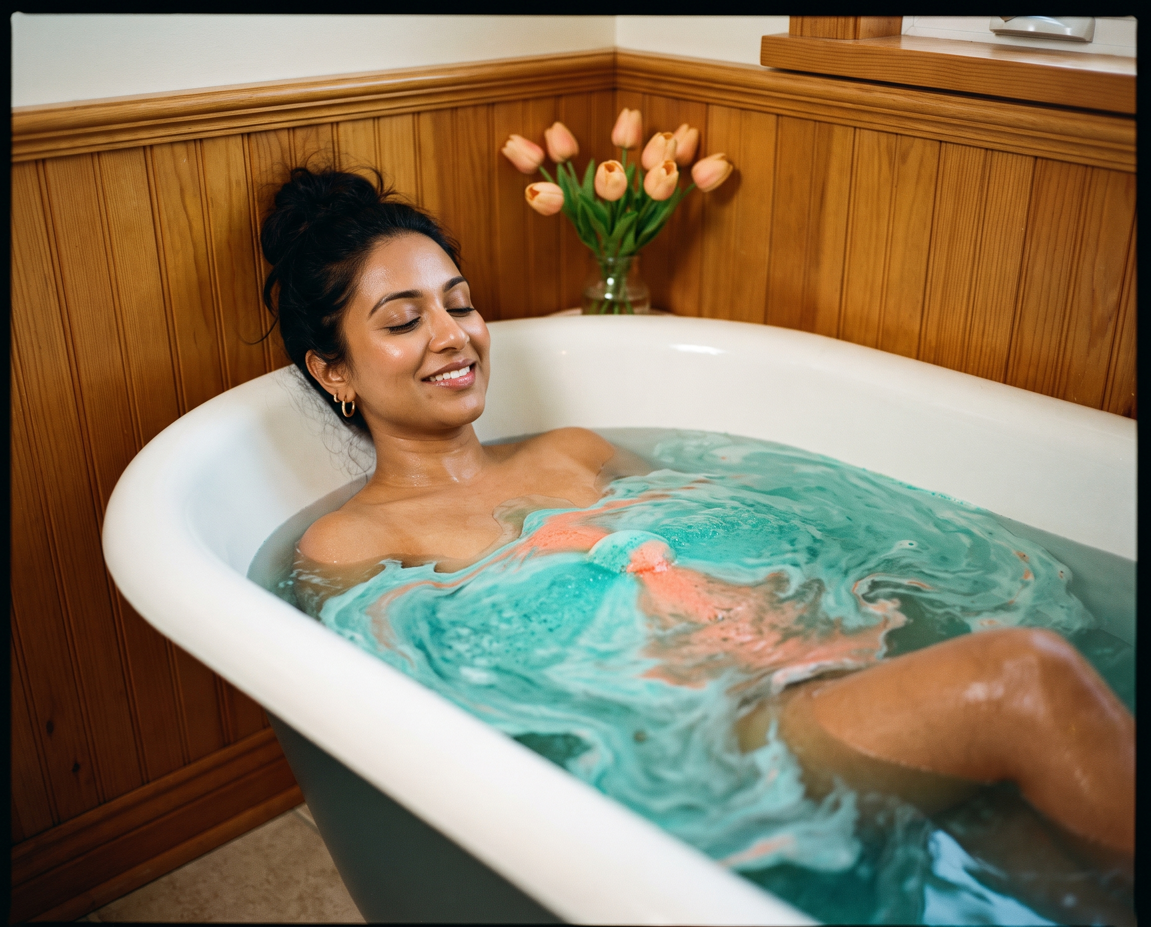 South Asian woman relaxing in a bathtub filled with swirling teal and coral water from a dissolving bath bomb
