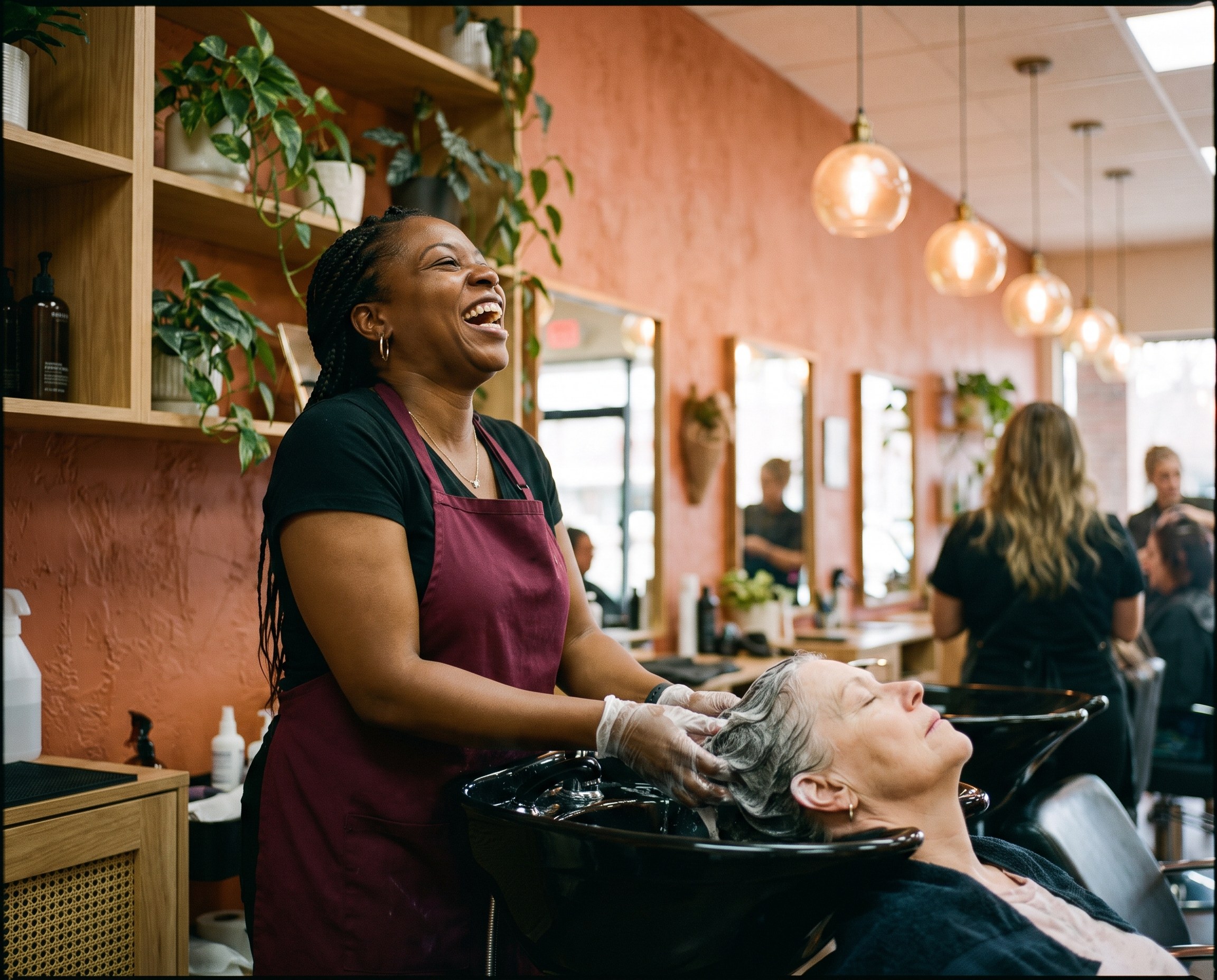 Stylist laughing while rinsing a client's gray hair at a salon basin