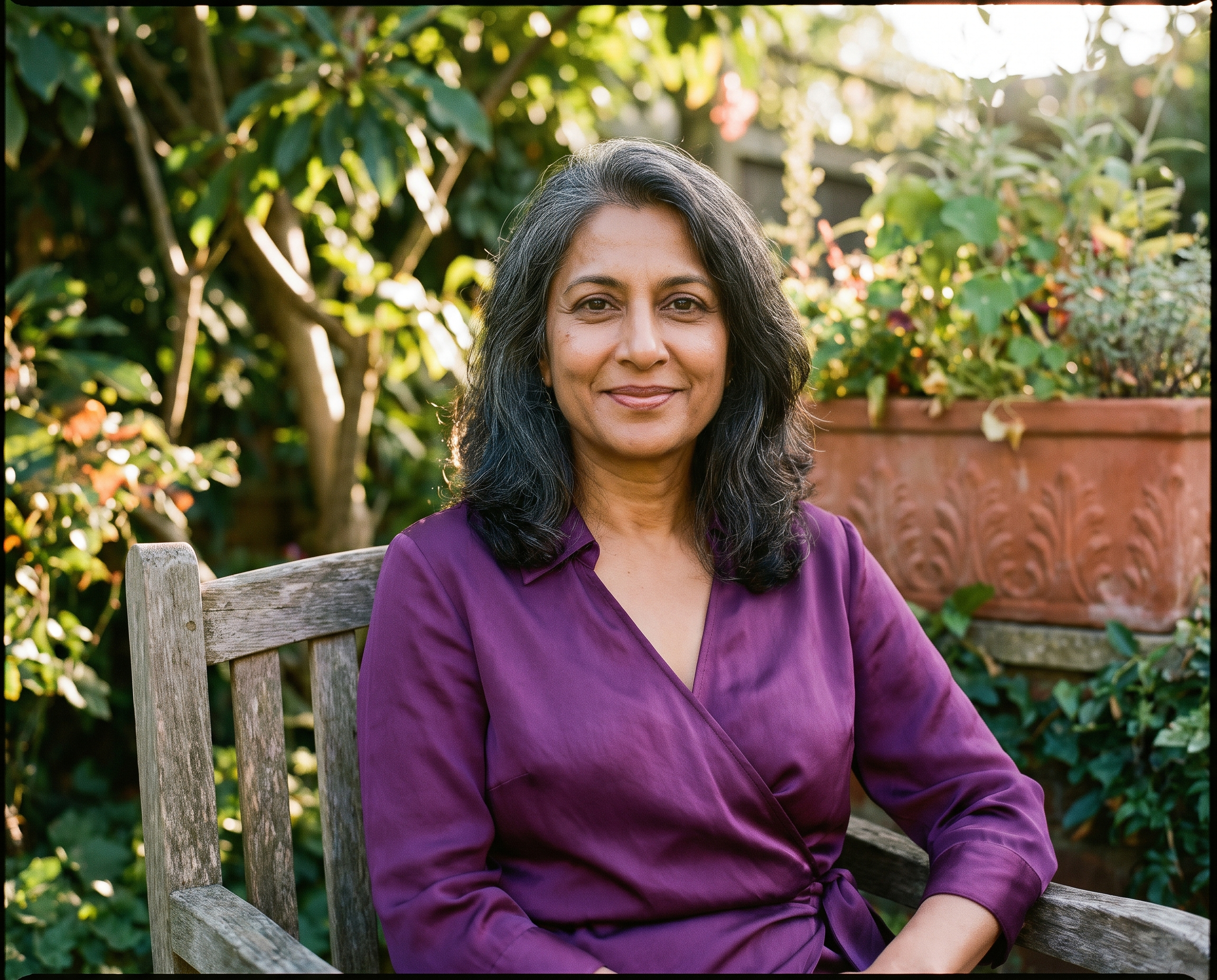 South Asian woman in her mid-50s seated outdoors in a garden chair beside green foliage, wearing an amethyst blouse