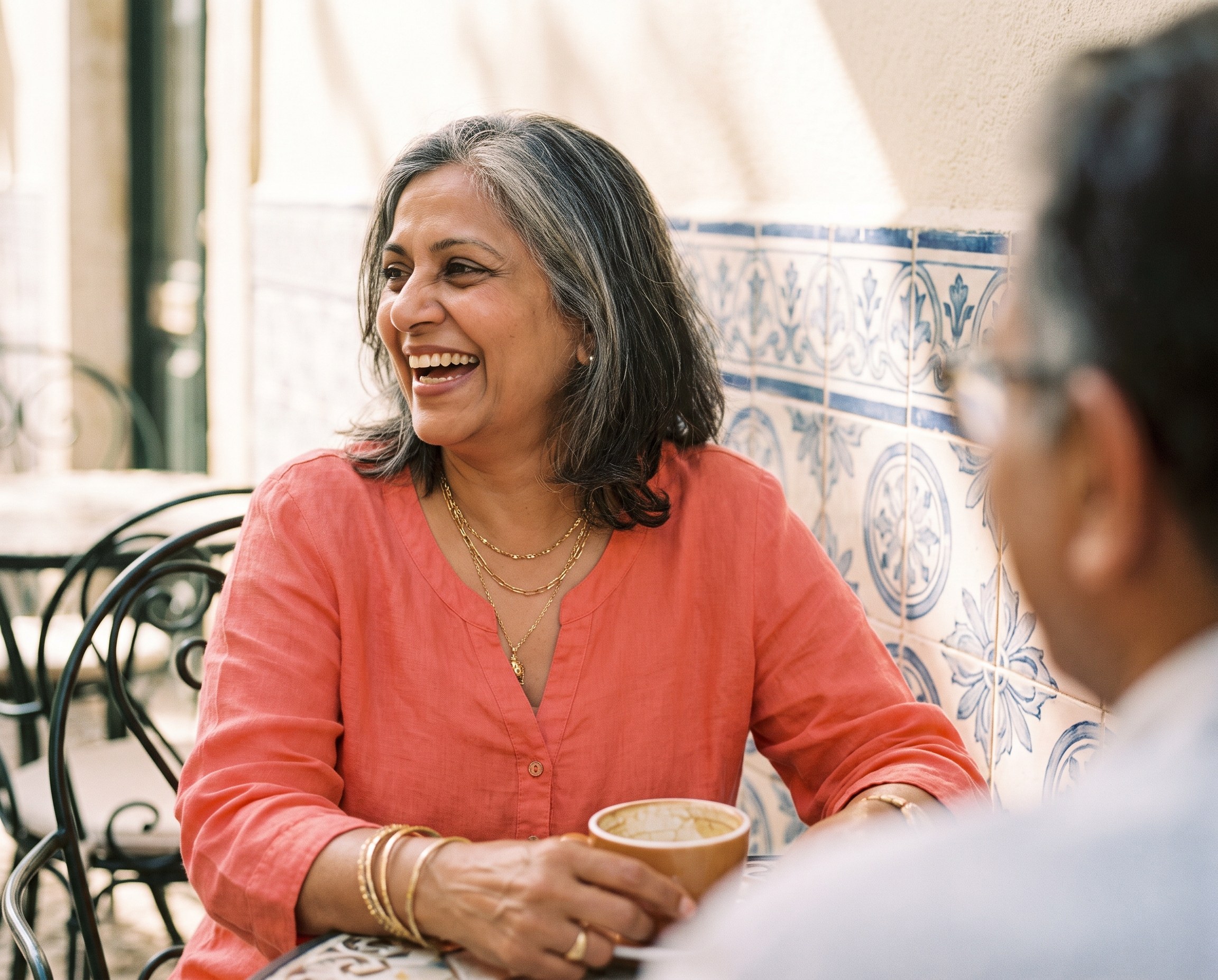 A radiant woman in her late 50s laughing at an outdoor cafe table with a colorful tiled wall behind her
