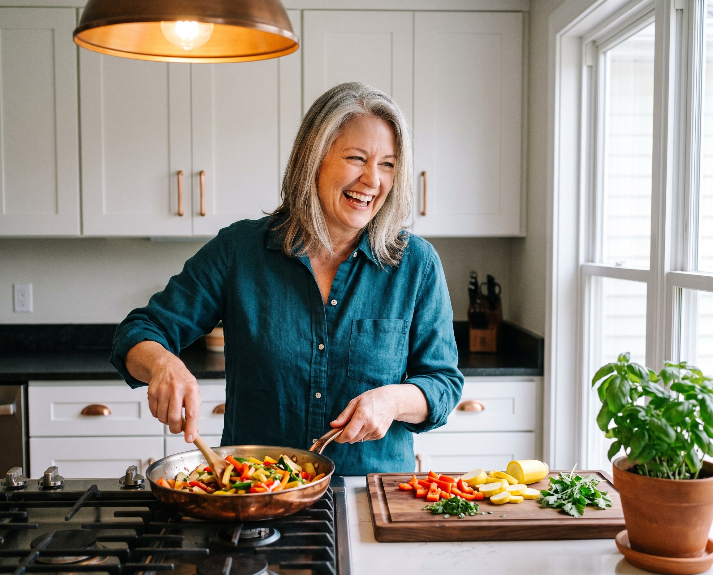 Silver-haired woman in a deep teal shirt preparing a colorful stir-fry in a bright modern kitchen with copper accents