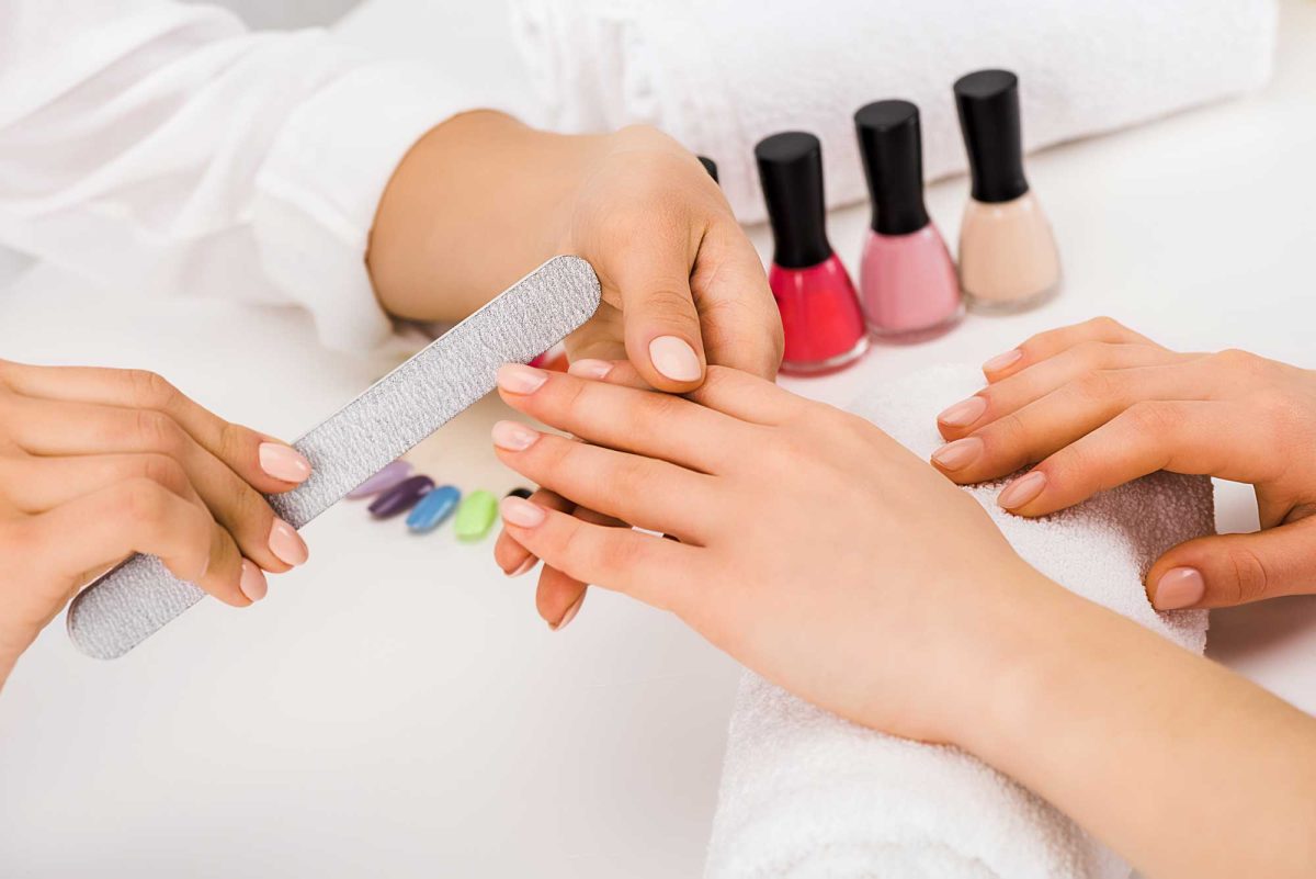 Manicurist files client’s nails beside colorful polish bottles.