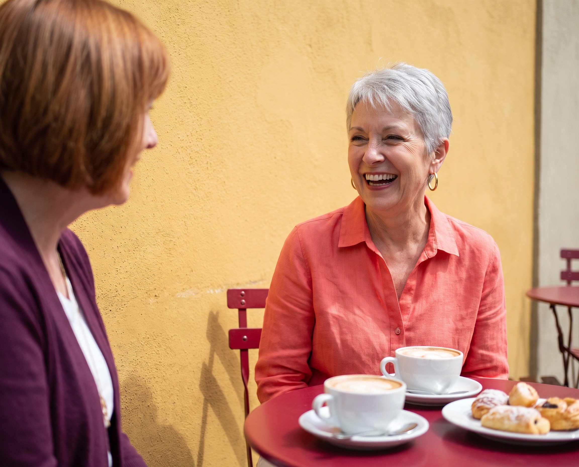 Woman around 60 with a silver pixie cut laughing at an outdoor cafe table