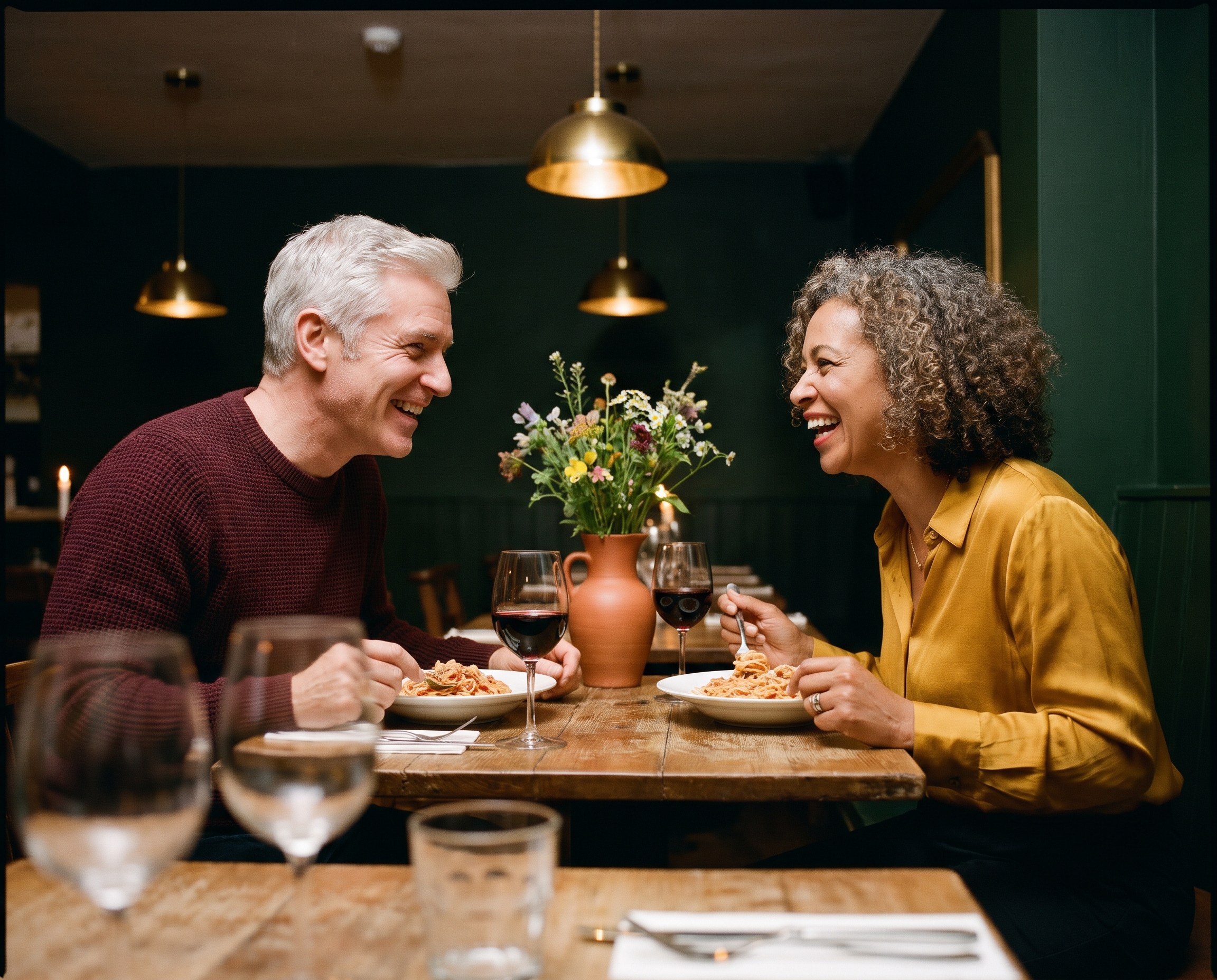 Two friends in their 60s laughing over dinner and red wine at a warmly lit restaurant