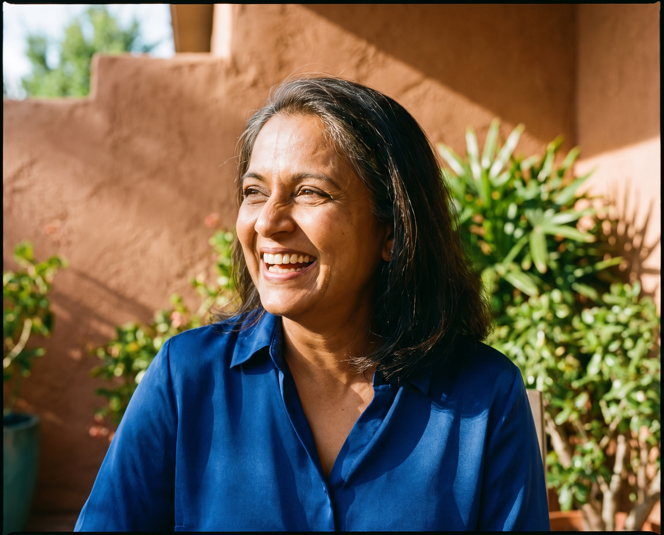 South Asian woman in her early 50s with golden-brown skin in a sapphire blue blouse laughing outdoors on a terracotta-walled patio