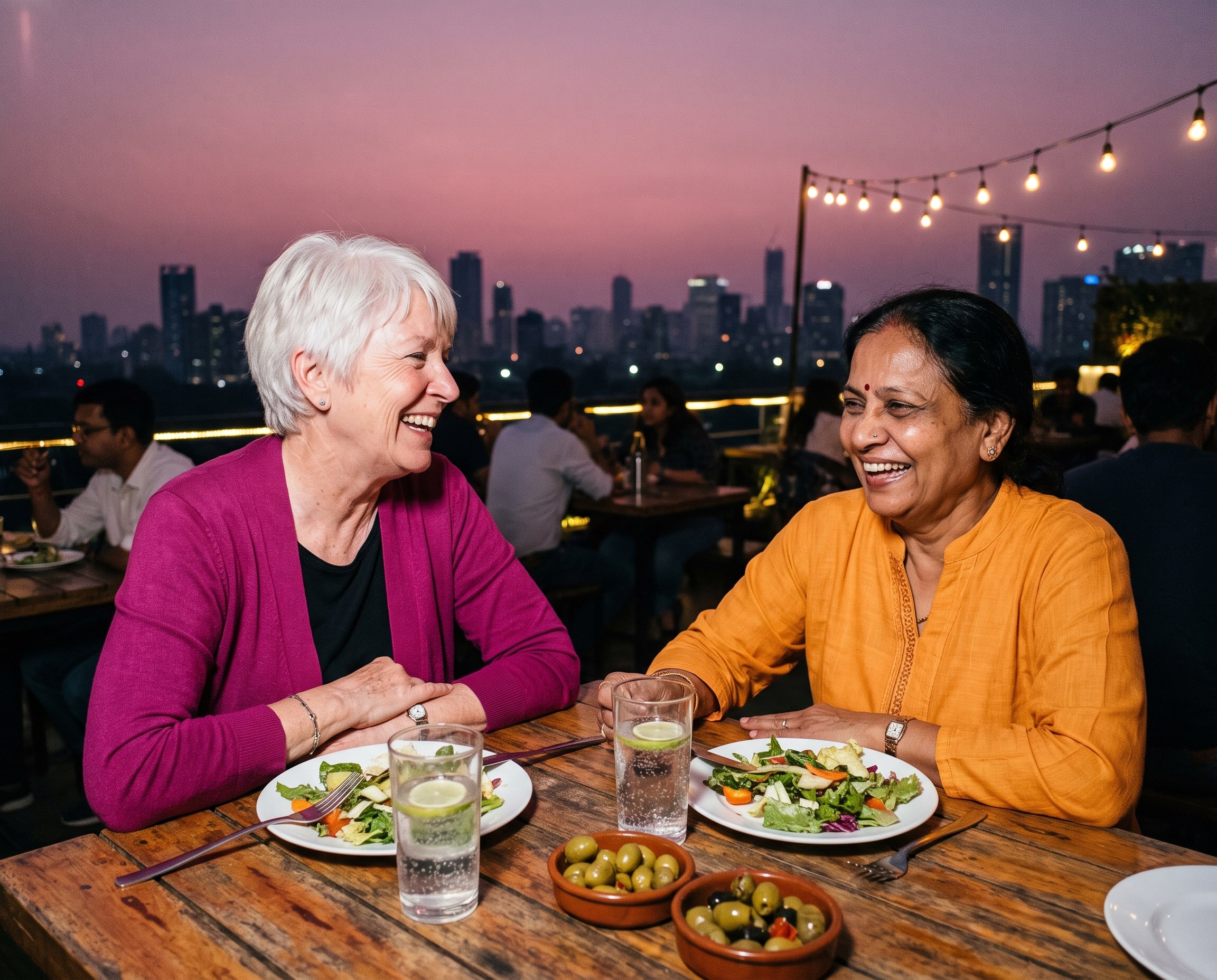 Two older women laughing over lunch at a rooftop table at dusk
