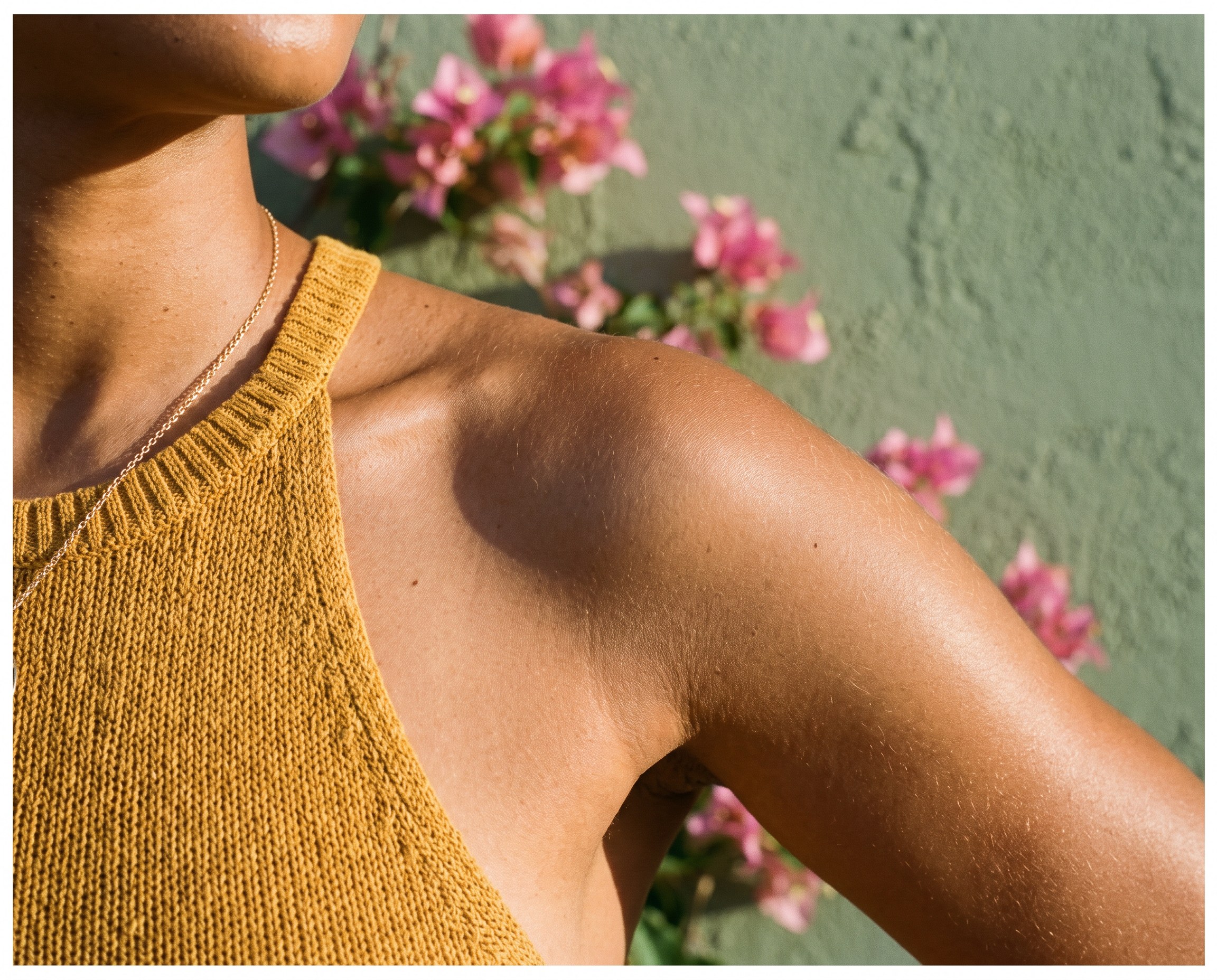 Close-up of sunlit shoulder and collarbone skin against a sage wall with bougainvillea