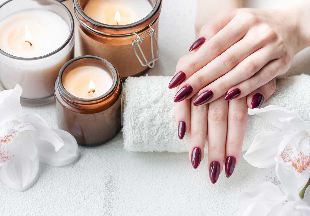 Burgundy manicured hands beside candles, towels, and white orchids
